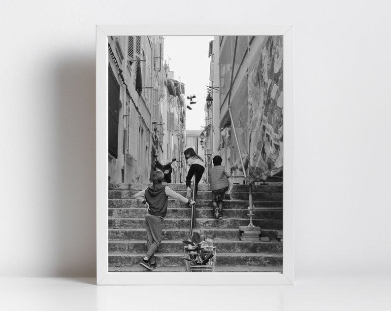 May include: Black and white framed photograph of children climbing a long set of outdoor stairs in a narrow European street. One child is leaning on a metal railing, while two others are ahead, ascending the steps. The scene is captured in natural light.