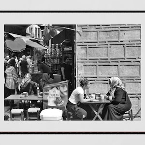 May include: Black and white photograph of a street scene with people. Two women sit at a table, while others are in the background near a shop with a Coca-Cola sign and Turkish flag.