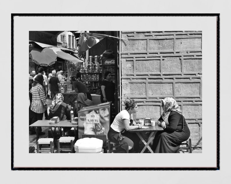 May include: Black and white photograph of a street scene with people. Two women sit at a table, while others are in the background near a shop with a Coca-Cola sign and Turkish flag.