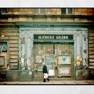 May include: A weathered storefront with a sign that reads "Uličnick&aacute; Galerie" in a faded blue and white striped awning. The storefront is covered in peeling paint and graffiti. A man in a dark jacket and pants walks past the storefront.