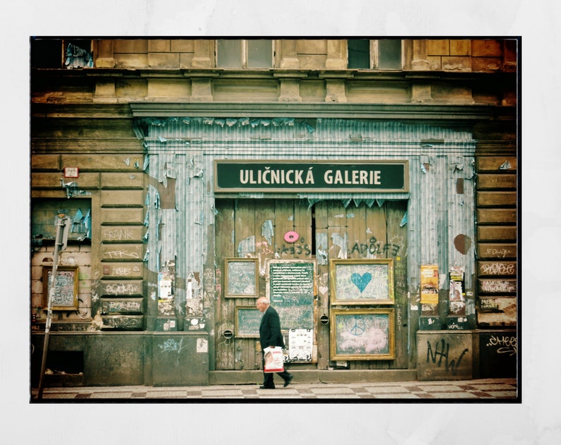May include: A weathered storefront with a sign that reads "Uličnick&aacute; Galerie" in a faded blue and white striped awning. The storefront is covered in peeling paint and graffiti. A man in a dark jacket and pants walks past the storefront.