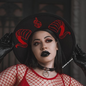 May include: A woman wearing a black wide-brimmed hat with red horns and flames, a red lace-up top with a fishnet overlay, and a black choker with silver rings. The hat has the text "Blessed by Lilith" on the side.