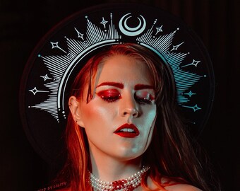 May include: A woman wearing a black dress and a red jacket with a star and moon crown pours a red liquid from a glass bottle into a cauldron. Green smoke rises from the cauldron.