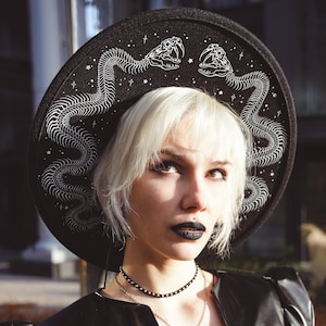 May include: A woman with blonde hair is wearing a black hat with a white snake design. The hat has a wide brim and is decorated with stars. The woman is also wearing a black leather jacket and a silver necklace.
