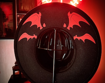 May include: A black wide-brimmed hat with red bat wings on the brim. The hat is on a black stand with a red base. The text "BLESSEDBYVILLAIN" is on the base of the stand.