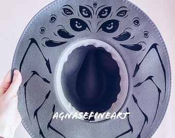 Könnte beinhalten: Grauer Filzhut im Fedora-Stil mit einem schwarzen Spinnenmotiv und mehreren schwarzen Augen auf der Krone. Der Hut trägt den Text "AGNASEFINEART" auf der Krempe.