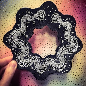 May include: Black embroidered patch featuring two snake skeletons in a circular design. The snakes are white, with detailed rib cages and skulls, set against a black background with white stars. The patch has a scalloped edge.