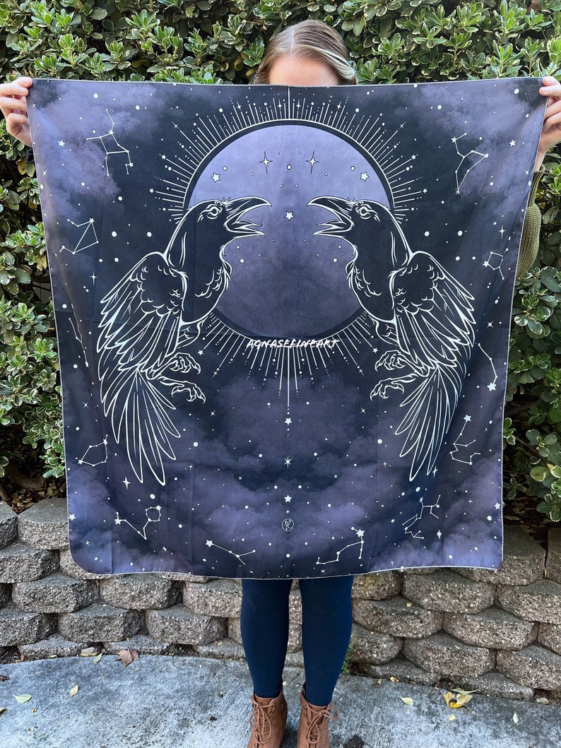 May include: A black tapestry featuring a white design of two ravens facing each other in front of a full moon with stars. The moon is encircled by a circle of rays. The background is a dark blue with white constellations.