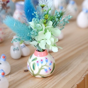 May include: A miniature ceramic vase with a pink rim and a floral pattern, containing a small bouquet of dried flowers in blue, green, and white. The vase is decorated with green vines and blue flowers. Other ceramic ornaments are in the background.