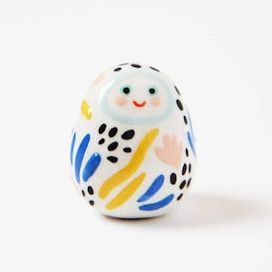 Op de afbeelding: Een wit keramiek beeldje met een glimlachend gezicht, blauwe ogen en een lichtblauwe wolkvorm op zijn hoofd. Het beeldje is versierd met zwarte, gele, blauwe en roze abstracte vormen.