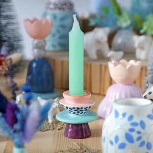 May include: A decorative candle holder with a mint green candle. The holder is composed of stacked ceramic elements in pink, blue, and purple. Other ceramic decor includes a blue vase, a pink tulip-shaped holder, and white bear figurines.