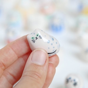 Peut inclure: Une petite figurine de chat en céramique blanche avec des pois bleus et des yeux verts. Le chat est assis avec sa queue enroulée autour de son corps.