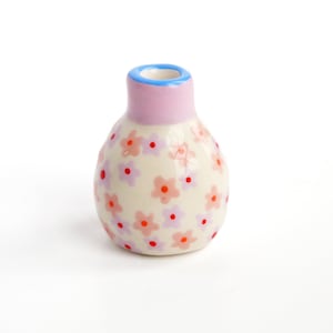May include: A small, white ceramic vase with a pink and blue rim. The vase is decorated with a floral pattern of pink and orange flowers with red centers.