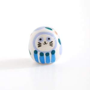 Puede incluir: Una pequeña figurita de cerámica blanca de un gato con detalles azules, verdes y naranjas. El gato tiene una cara sonriente y lleva un sombrero a rayas azul y verde.
