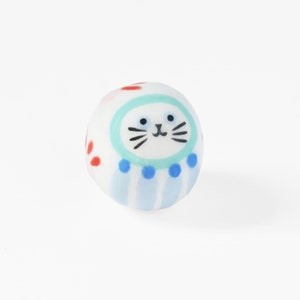 Peut inclure: Une boule ronde en céramique blanche avec un motif rayé bleu et blanc, des points rouges et un visage de chat bleu et blanc avec des moustaches noires.