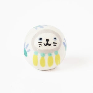 Puede incluir: Una figurita de cerámica blanca de un gato con la cara gris, detalles florales azules y verdes y flecos amarillos y verdes.