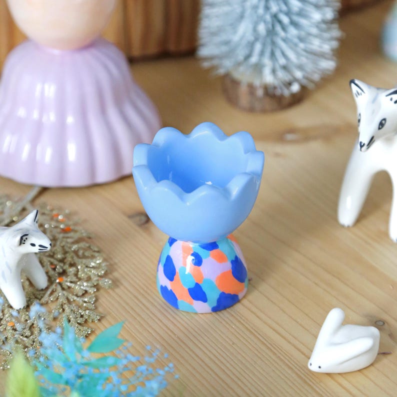 May include: A light blue ceramic egg cup with a colorful, abstract base. The egg cup is surrounded by small decorative figurines, including a white fox and a white rabbit, on a wooden surface. Other decorative items are in the background.