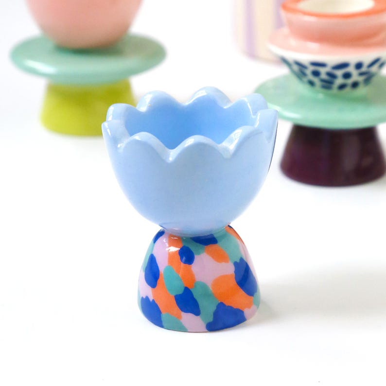 May include: A collection of colorful ceramic egg cups. The front-most cup is light blue with a scalloped rim, atop a base with a colorful abstract pattern of blue, orange, and pink. Other cups in the background are pink, green, and purple.
