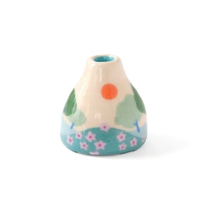 May include: A small ceramic vase with a white background and a painted scene of a green landscape with a blue sky, pink flowers, and an orange sun.