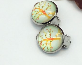 May include: A pair of silver clip-on earrings with a round, light blue background and a yellow tree with pink flowers.