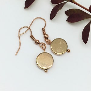 Puede incluir: Un par de pendientes de color dorado con dijes redondos en forma de disco. Los pendientes tienen un diseño simple y minimalista.