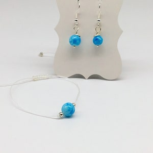 Könnte beinhalten: Ein Set aus blauen Agat-Ohrringen und einem passenden Armband. Die Ohrringe sind silberfarben mit einem blauen Agatsteine. Das Armband ist ein weißes Band mit einem blauen Agatsteine.