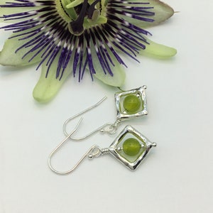 Op de afbeelding: Zilveren oorbellen met een vierkant frame en een groene edelsteen in het midden. De oorbellen hangen aan een zilveren haak.