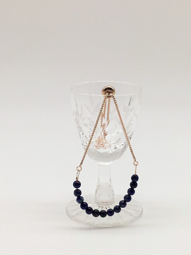 May include: A gold chain bracelet with a row of dark blue beads. The bracelet is displayed on a clear glass pedestal.