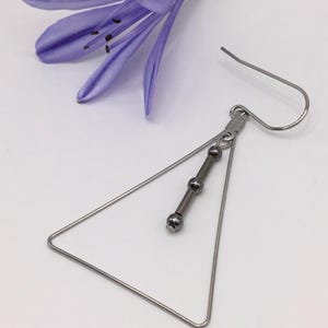 Può includere: Orecchino geometrico color argento con una cornice triangolare e una barra verticale con tre perline scure. L'orecchino è mostrato con un fiore viola su sfondo bianco. Chiusura a gancio.