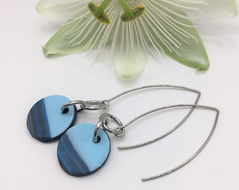 Puede incluir: Pendientes colgantes con colgantes redondos veteados en azul y negro. Los pendientes tienen alambres para las orejas curvados de color plateado. Una flor blanca está en el fondo. Un accesorio elegante.