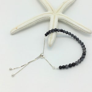 May include: A black and grey gemstone bracelet with a silver adjustable sliding clasp. The bracelet is made of small round beads and is photographed on a white background with a white starfish.