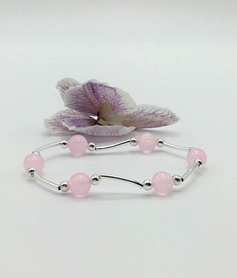 May include: A delicate bracelet with a silver chain and pink beads. The bracelet is displayed on a white background with a dried orchid flower.
