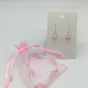 May include: A set of pink gemstone earrings and a bracelet. The earrings are silver with a pink gemstone dangle. The bracelet is silver with pink gemstones and is in a pink organza bag.