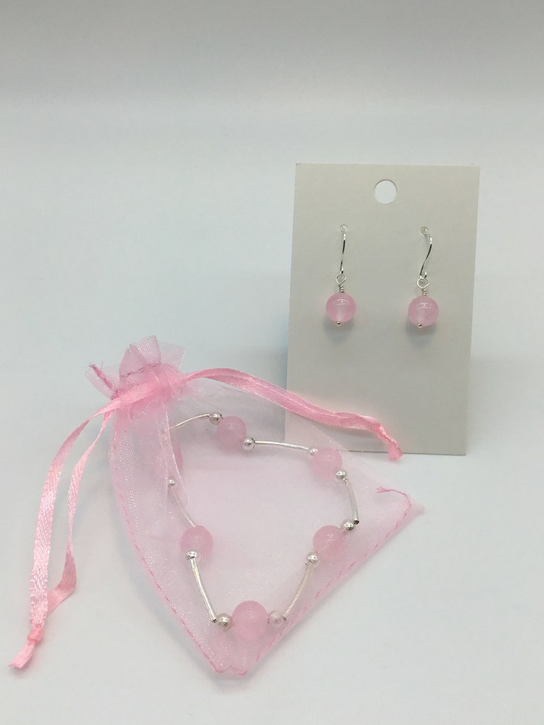 May include: A set of pink gemstone earrings and a bracelet. The earrings are silver with a pink gemstone dangle. The bracelet is silver with pink gemstones and is in a pink organza bag.