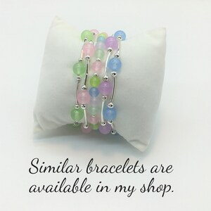 May include: A set of five delicate bracelets with pastel-colored beads and silver accents. The bracelets are layered on a white surface.