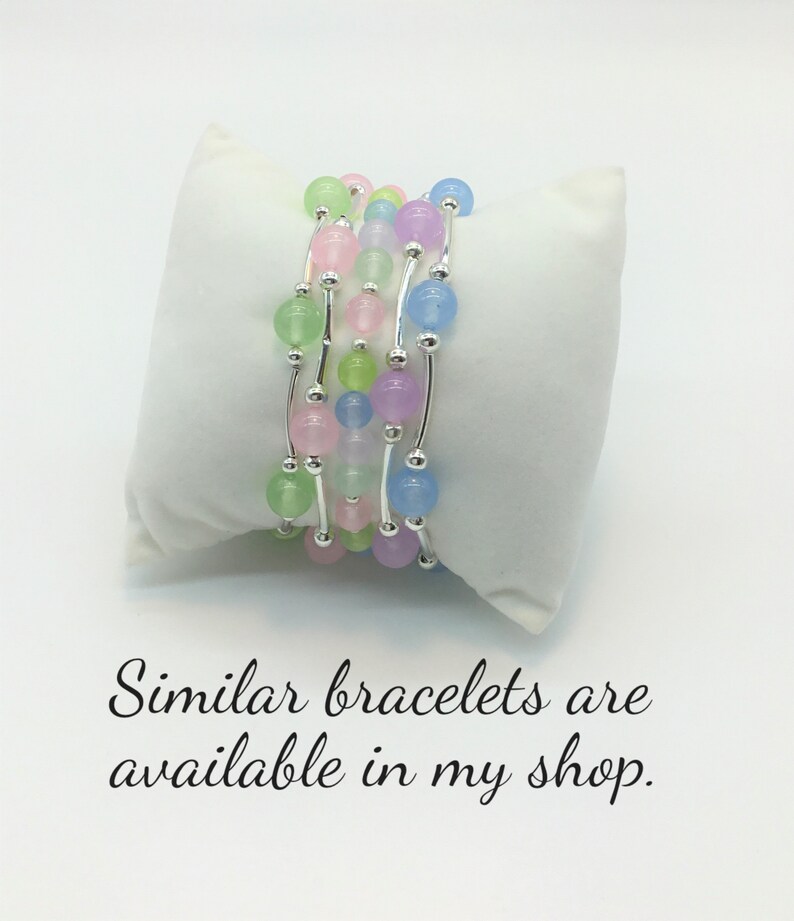 May include: A set of five delicate bracelets with pastel-colored beads and silver accents. The bracelets are layered on a white surface.