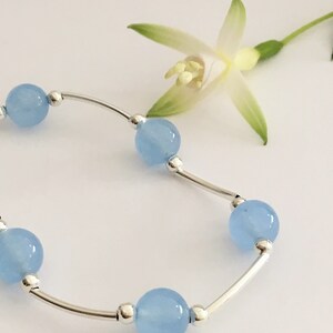 May include: A delicate silver bracelet with light blue round beads. The bracelet is on a white background with a white flower.
