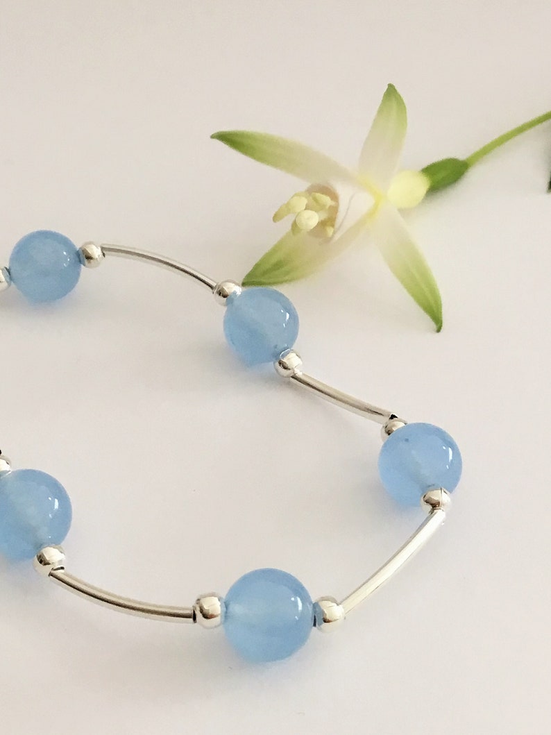 May include: A delicate silver bracelet with light blue round beads. The bracelet is on a white background with a white flower.