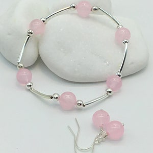 May include: A set of pink gemstone jewelry featuring a bracelet and earrings. The bracelet is made of silver and has pink round beads. The earrings are silver with pink round beads.