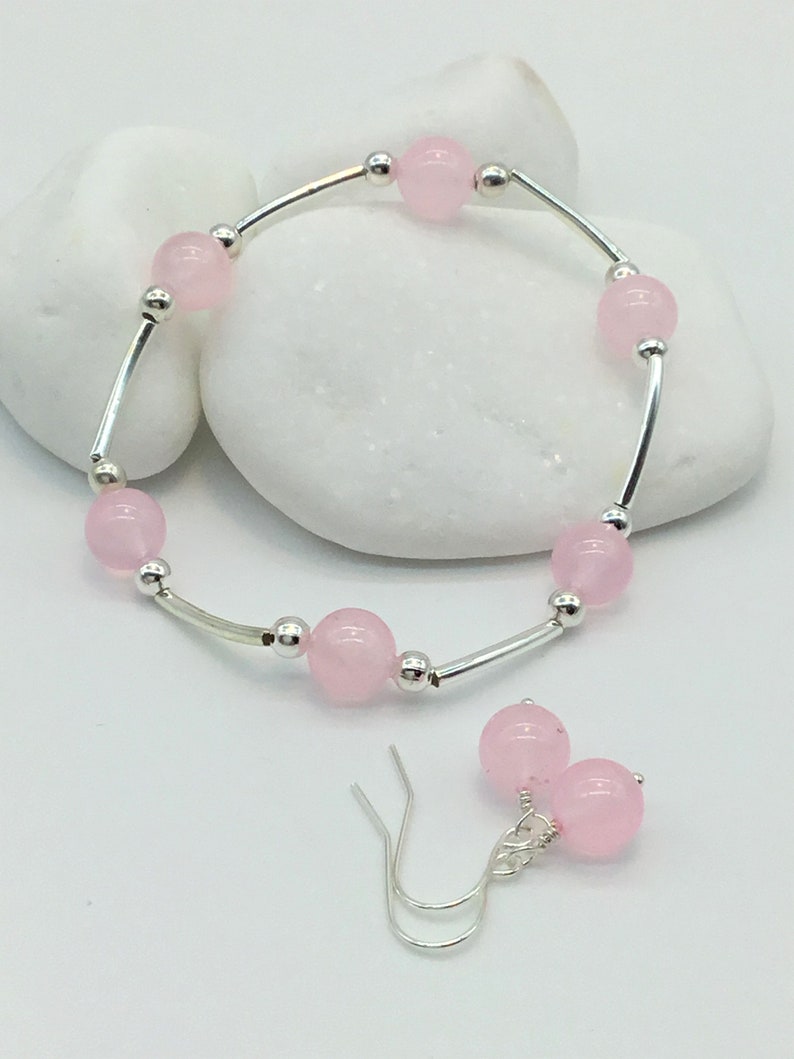 May include: A set of pink gemstone jewelry featuring a bracelet and earrings. The bracelet is made of silver and has pink round beads. The earrings are silver with pink round beads.