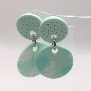 Peut inclure: Une paire de boucles d'oreilles pendantes vert menthe avec un motif géométrique blanc et bleu clair. Les boucles d'oreilles sont en pâte polymère et ont des crochets argentés.