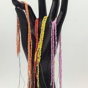 Op de afbeelding: Een set van zes kralenkettingen in verschillende kleuren, waaronder oranje, geel en roze. De kettingen zijn geregen aan dunne zilveren koorden en worden getoond op een zwarte handvormige standaard.