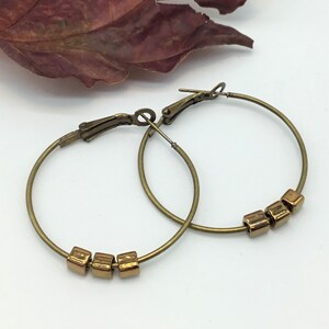 May include: A pair of gold hoop earrings with small gold square beads. The earrings are made of metal and have a simple design.