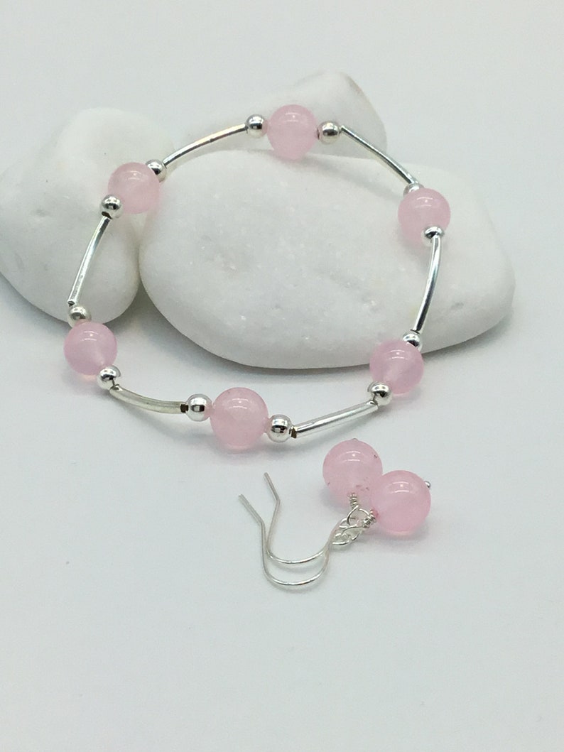 May include: A set of pink gemstone jewelry featuring a bracelet and earrings. The bracelet is made of silver and has pink round beads. The earrings are silver with pink round beads.