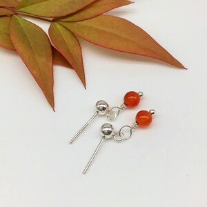 May include: A pair of silver stud earrings with a small orange carnelian bead dangling from each. The earrings are on a white background.