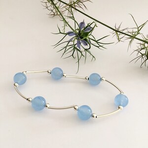 May include: A delicate silver bracelet with light blue round beads. The bracelet is on a white background with a flower.