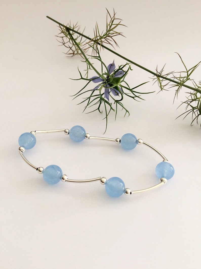 May include: A delicate silver bracelet with light blue round beads. The bracelet is on a white background with a flower.