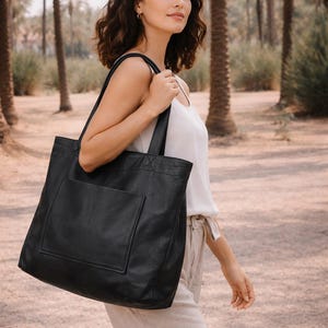 May include: A large black tote bag with a front pocket is carried over the shoulder. The bag is made of a smooth material and has a rectangular shape. The background features palm trees and a pathway.