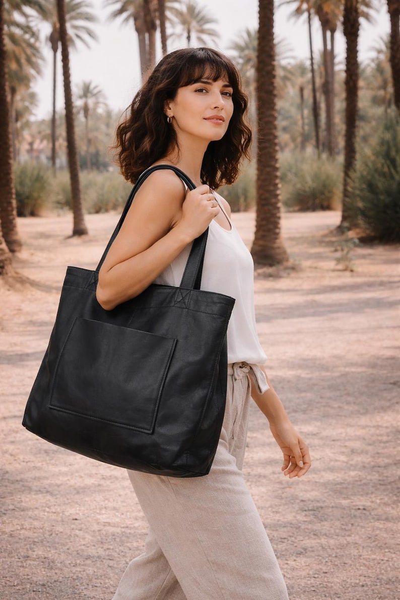 May include: A large black tote bag with a front pocket is carried over the shoulder. The bag is made of a smooth material and has a rectangular shape. The background features palm trees and a pathway.