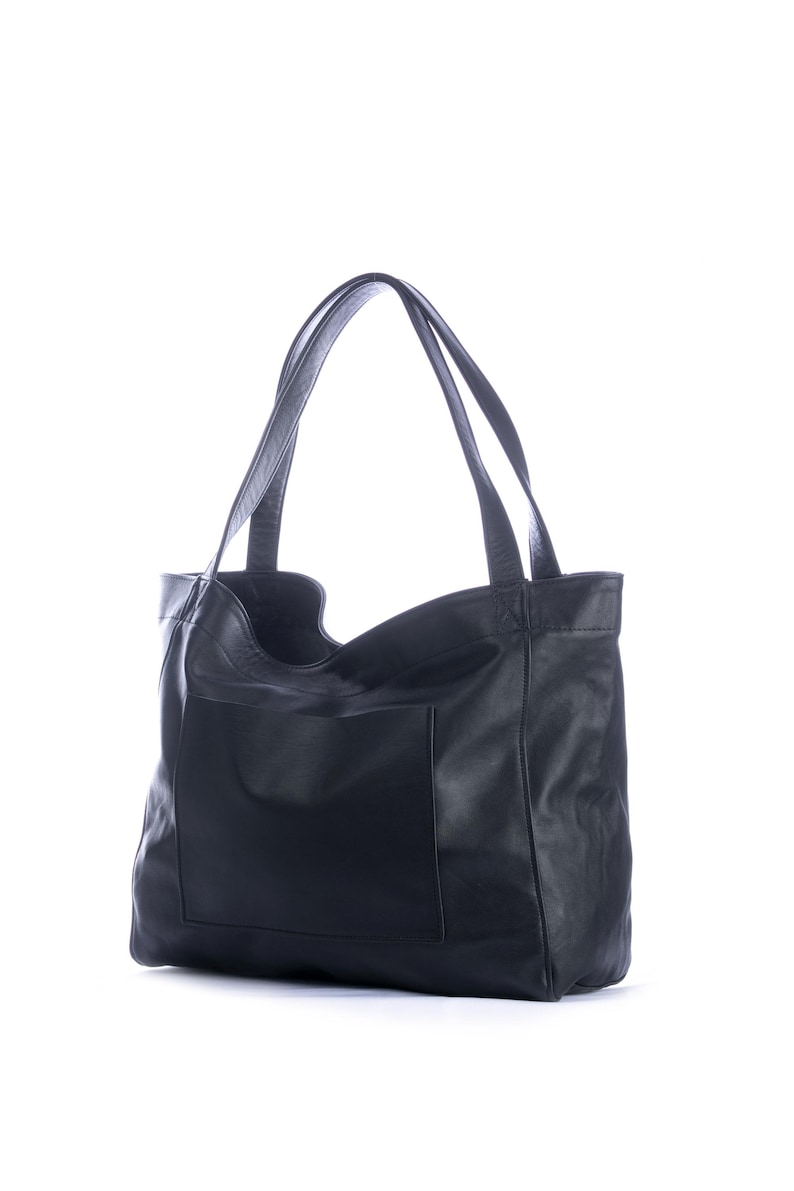 May include: A black leather tote bag with two long handles and a large front pocket. The bag has a simple, classic design and is made of a soft, textured leather. The bag is empty and stands upright against a white background.